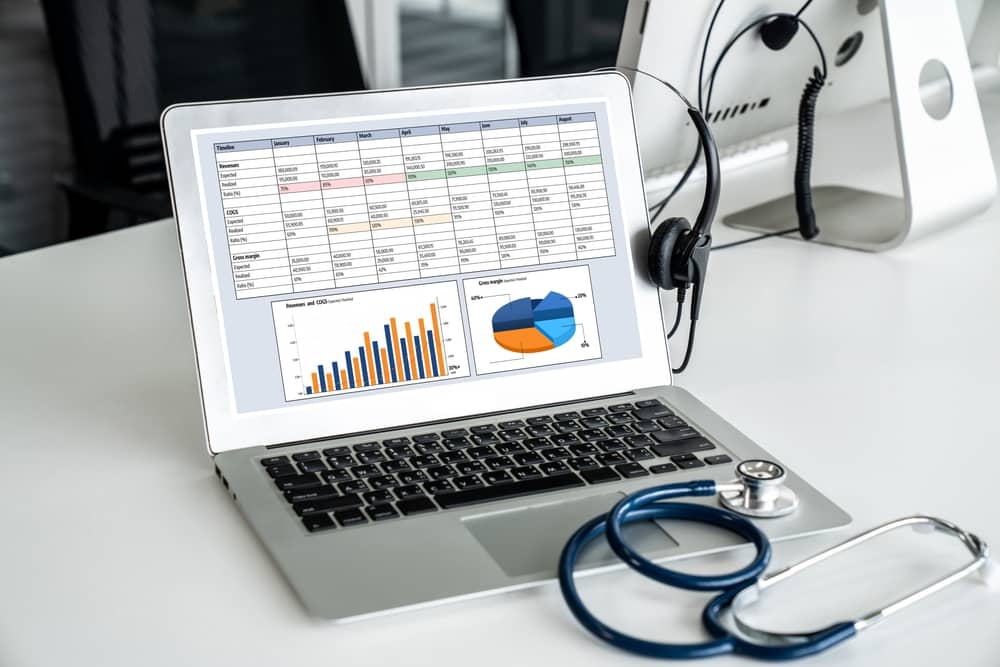 Laptop displaying healthcare analytics dashboard with spreadsheets, bar charts, and pie graphs next to stethoscope and medical equipment in clinical setting