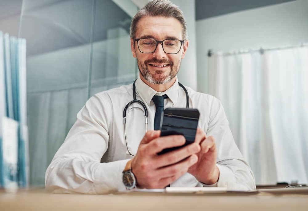Smiling male doctor with glasses and stethoscope using smartphone in modern medical clinic hallway
