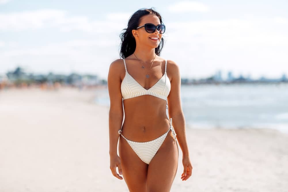 Confident woman in bikini on beach showing flat stomach and toned body after successful fat reduction treatment and body transformation