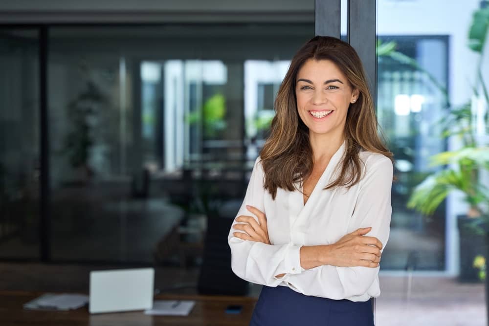 Confident plastic surgery copywriter standing in modern office, arms crossed with warm smile, representing professional expertise