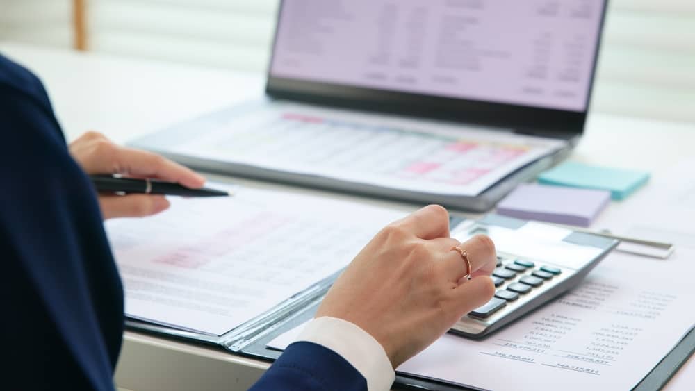 Business professional in navy suit using calculator and reviewing financial documents with laptop and charts on desk
