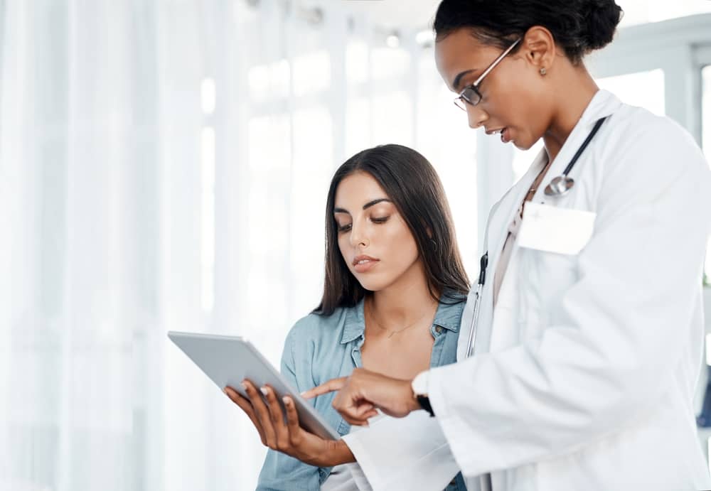 Female doctor with stethoscope showing tablet information to young woman patient in medical consultation, discussing plastic surgery insurance coverage options