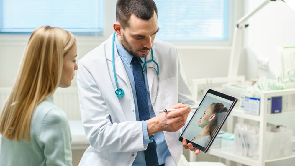 Professional male doctor in white coat with stethoscope showing tablet with aesthetic treatment results to blonde female patient during med spa consultation in modern medical office, demonstrating trust-building consultation process