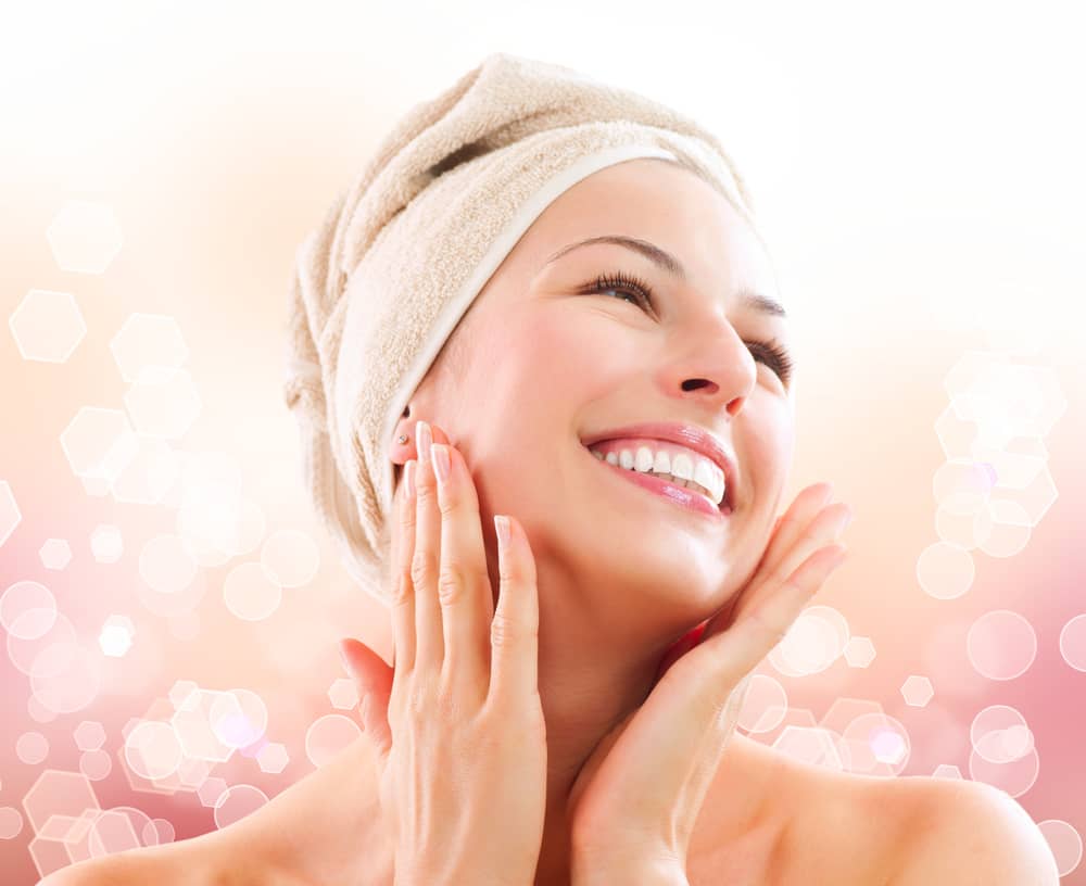 Radiant woman with glowing skin wearing spa headband and bright smile, touching her face with both hands in joy and confidence, representing successful aesthetic treatment results and emotional transformation on soft pink bokeh background