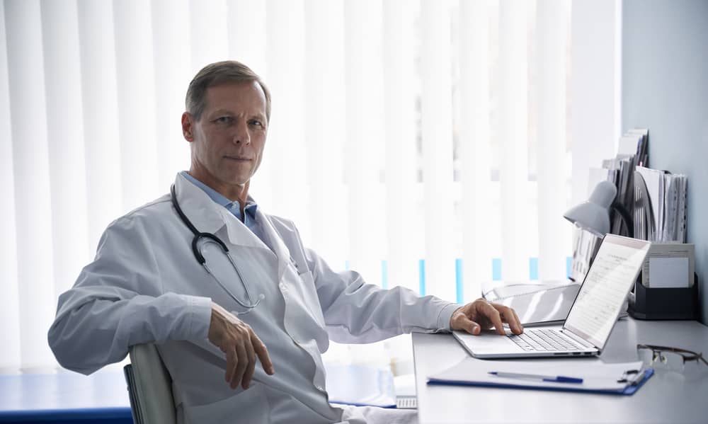 Confident middle-aged physician in white coat sitting at desk with laptop in bright modern office, representing the authoritative leadership and systematic control that elite surgeons maintain over their practice operations