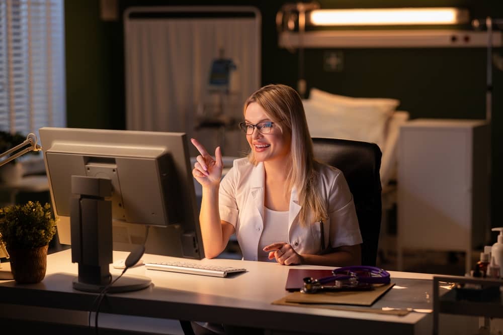 Blonde female healthcare professional in white coat pointing at computer screen while smiling in modern medical office, representing expert copywriting for plastic surgery practices