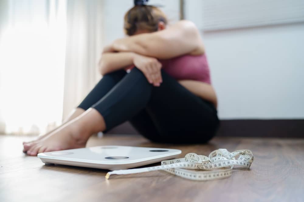 Frustrated woman sitting on floor next to scale and measuring tape representing the struggle with stubborn body fat that diet and exercise can't eliminate