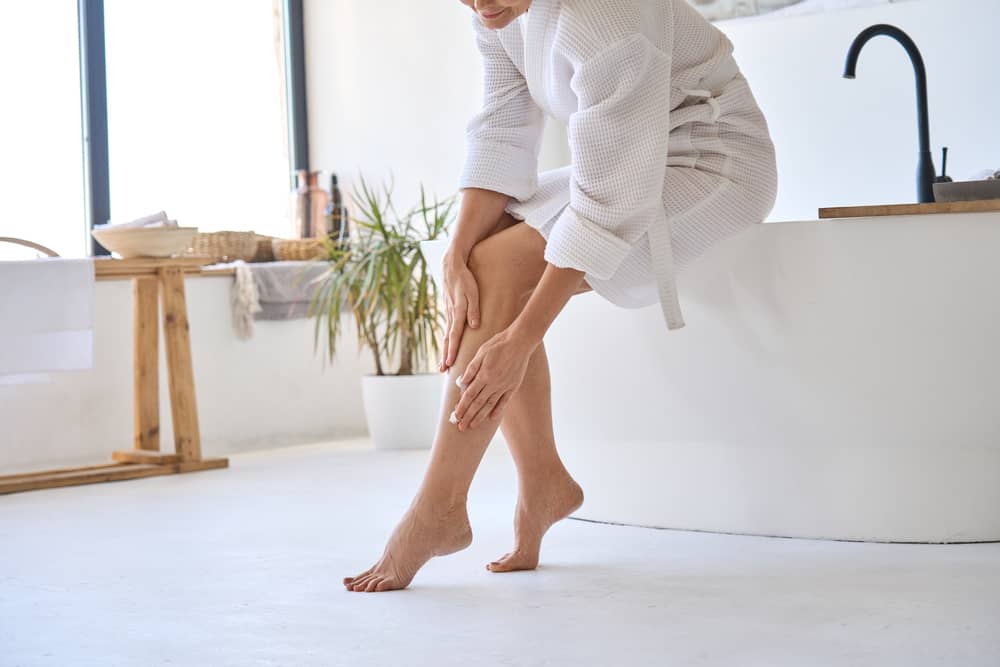 Woman in white robe applying moisturizer to legs after laser hair removal treatment in modern bathroom