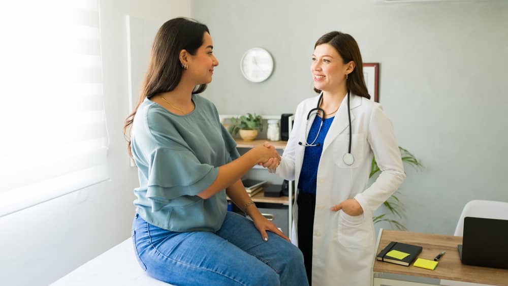 Smiling female doctor in white coat shaking hands with happy patient in blue sweater during successful plastic surgery consultation in modern medical office