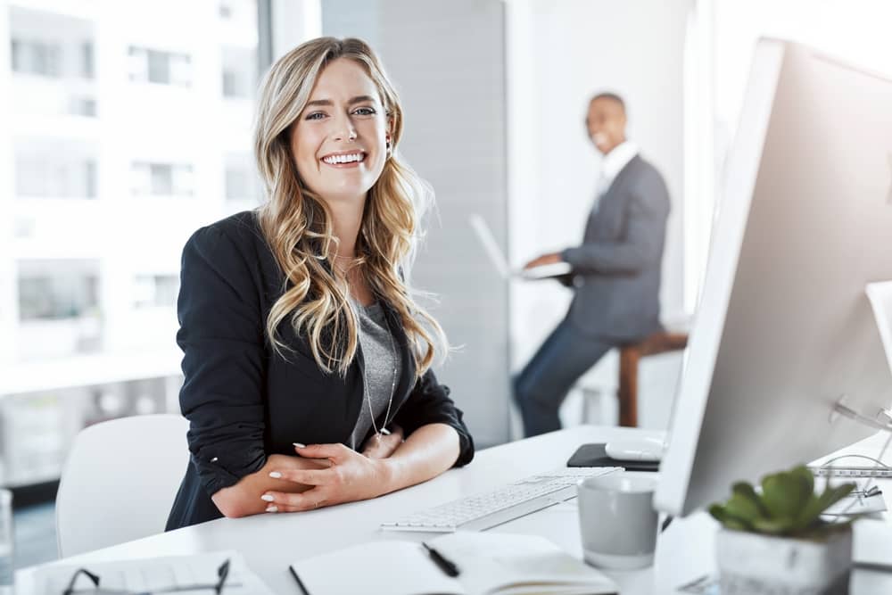 Confident blonde businesswoman in black blazer smiling at desk in modern office with male colleague working in background
