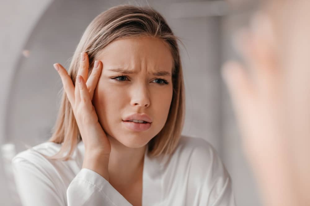 Young blonde woman looking worried while examining her reflection in a bathroom mirror, touching her face with concern about aging and skin appearance, perfect for med spa marketing about client insecurities and beauty concerns
