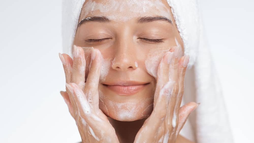 Woman gently cleansing face with soap and water, hands cupping cheeks with white foam cleanser