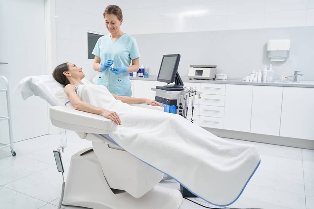 Smiling aesthetician consulting with relaxed client on treatment chair in modern med spa with HydraFacial equipment
