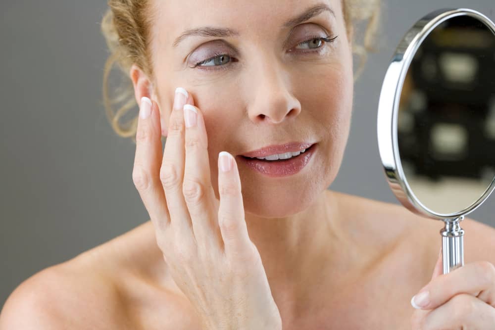 Mature woman examining her smooth, youthful skin in handheld mirror, gently touching her face with satisfied expression