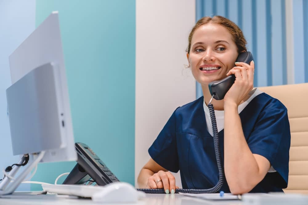 Smiling medical receptionist in navy scrubs speaking on the phone while sitting at a desk with a computer.