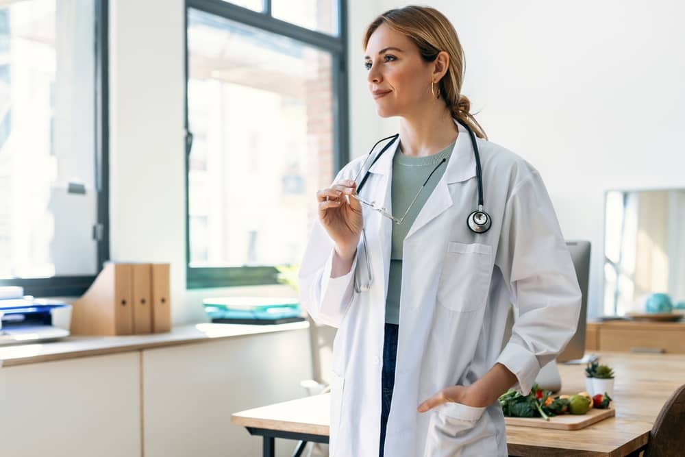 Confident female medical professional in white lab coat with stethoscope looking thoughtfully out window in bright modern medical office, representing trusted aesthetic practitioner expertise and patient care