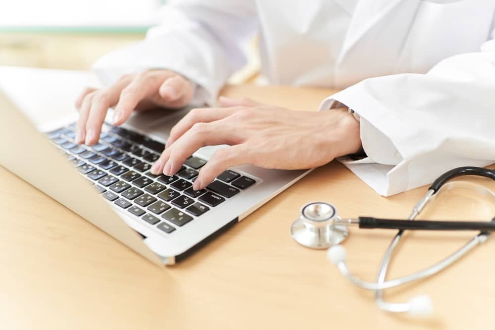 Healthcare professional in white coat typing on laptop with stethoscope on wooden desk