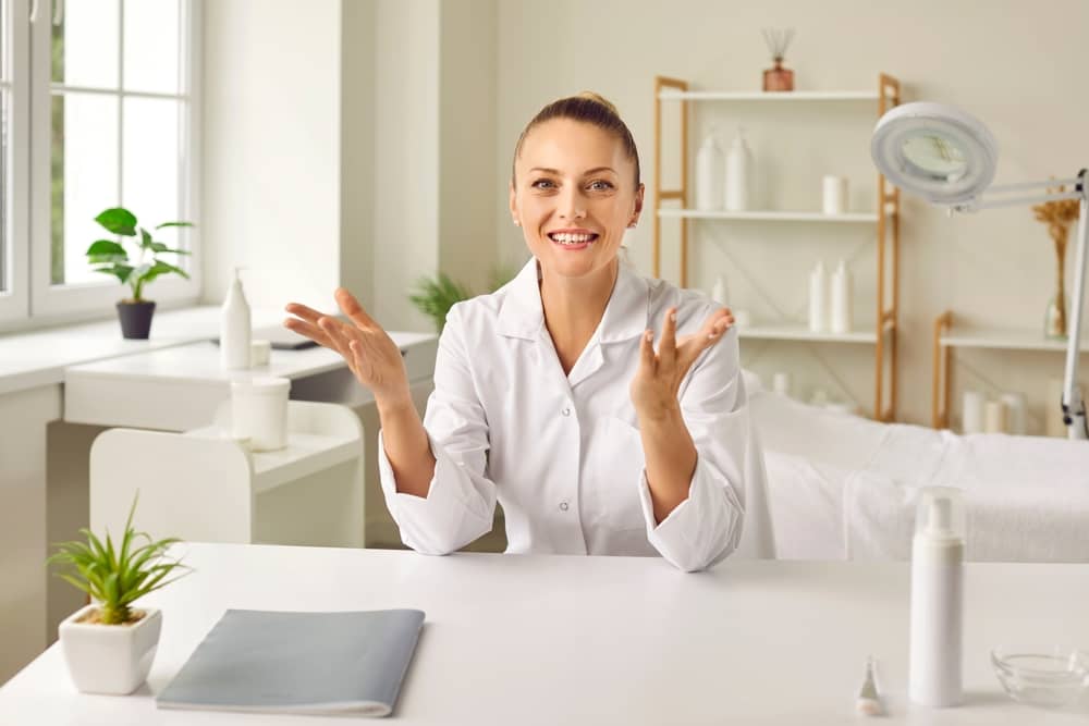 Smiling female aesthetic practitioner in white medical coat sitting at consultation desk in modern medical spa office, gesturing welcomingly with both hands raised in professional, friendly manner