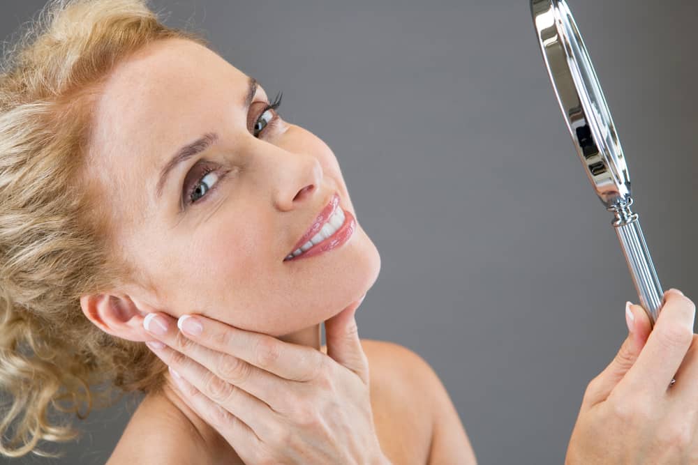 Smiling mature woman with blonde hair examining her firm jawline and neck in handheld mirror, demonstrating confidence in her youthful-looking skin after non-surgical aesthetic treatment