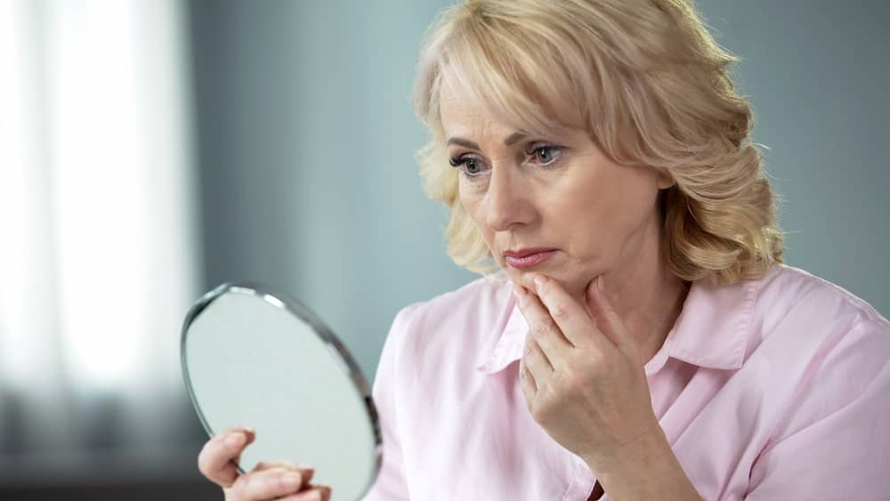 Concerned mature woman in pink shirt examining her face and neck in handheld mirror, touching her chin while looking worried about visible signs of aging and skin laxity