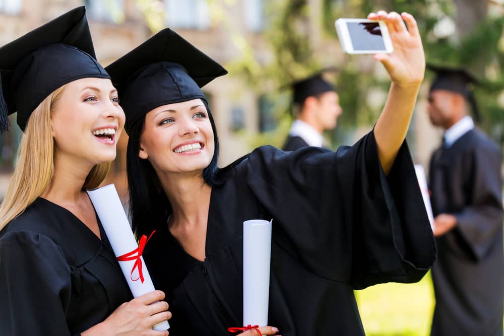 Two female graduates in black caps and gowns taking selfie with diplomas, representing med spa business owners who started with same education but achieved different success levels