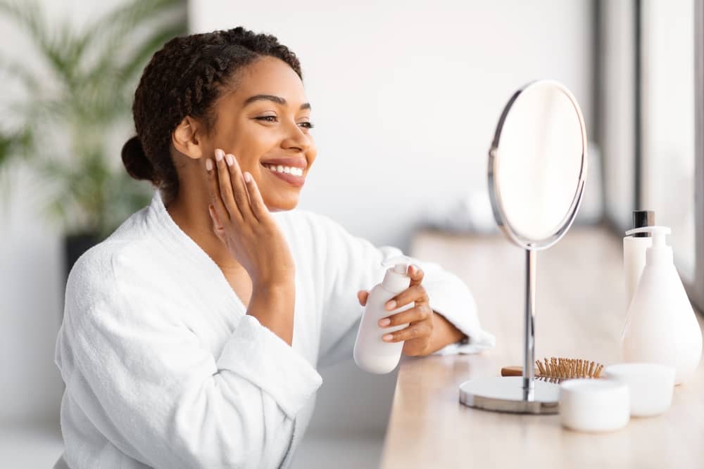 Smiling Black woman applying gentle moisturizer as part of aftercare routine following safe laser treatment on dark skin