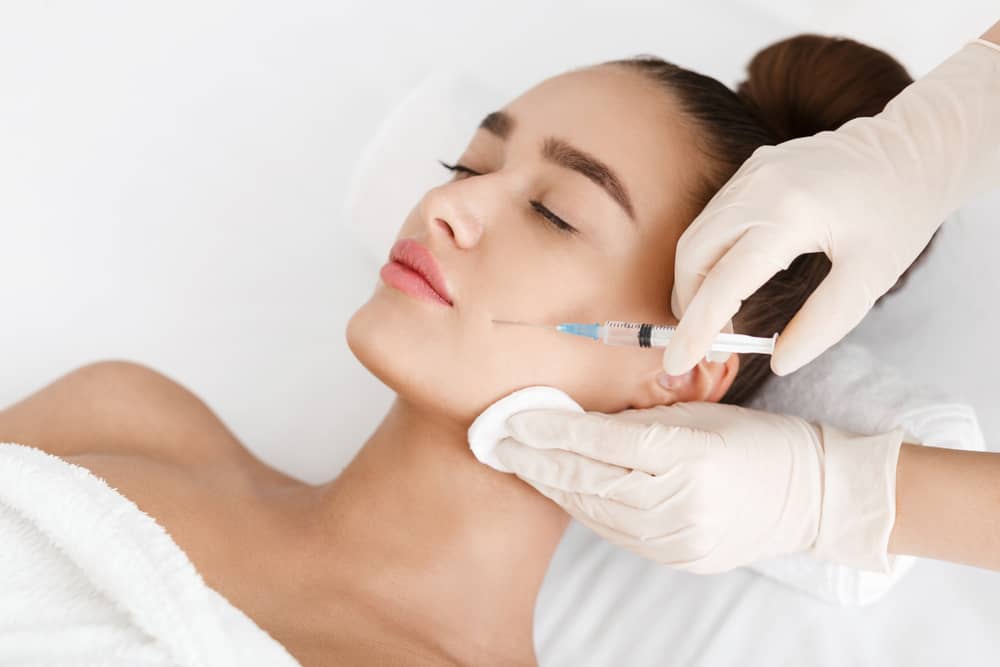 Aesthetic injector administering dermal filler treatment to young woman's jawline at medical spa