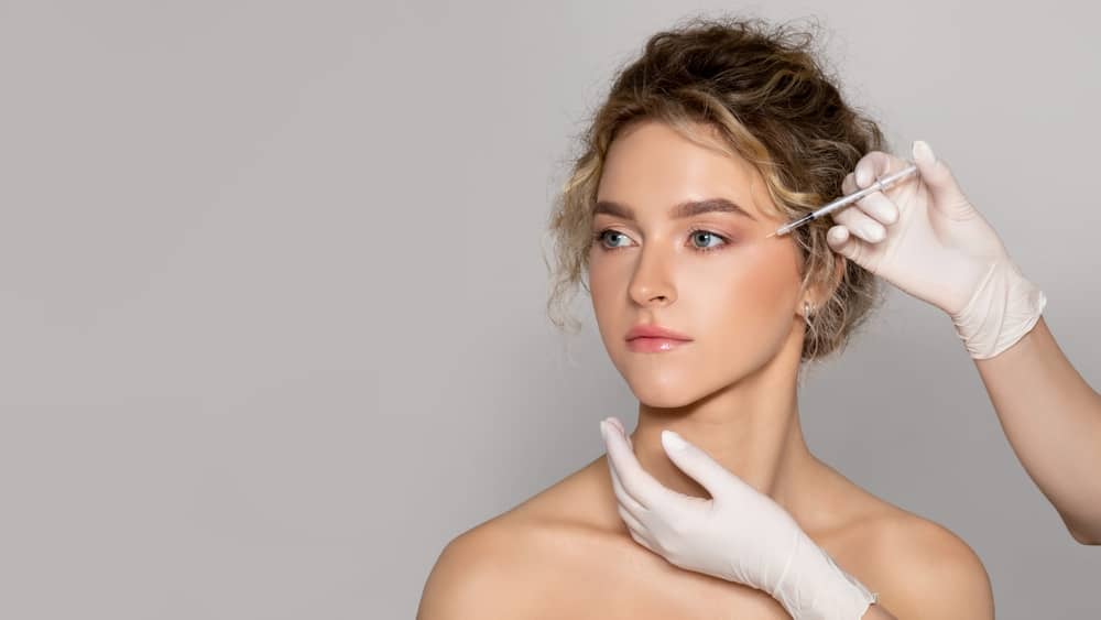 Young woman receiving botulinum toxin injection near eye area by aesthetics provider in white gloves