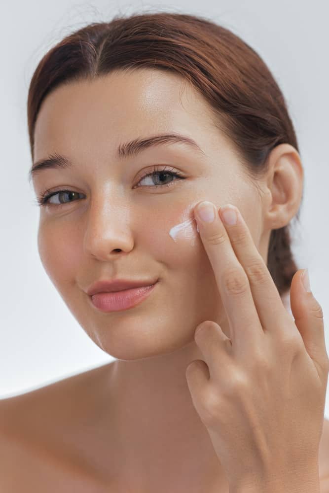 Young woman with smooth skin applying topical skincare cream to her cheek demonstrating evidence-based wrinkle prevention