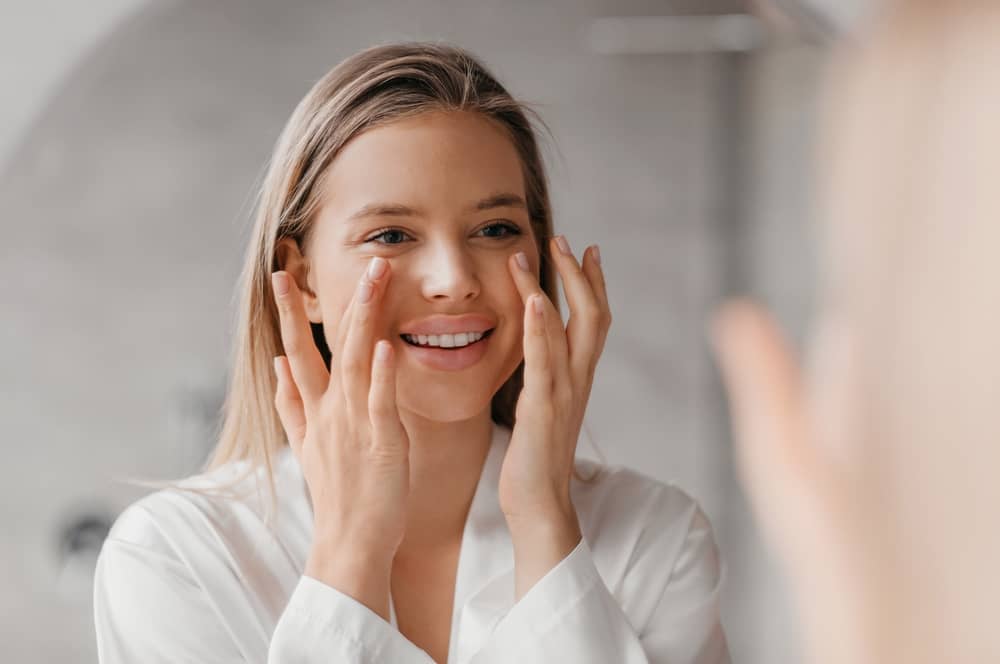 Young woman applying face cream looking in mirror smiling Gen Z skincare routine