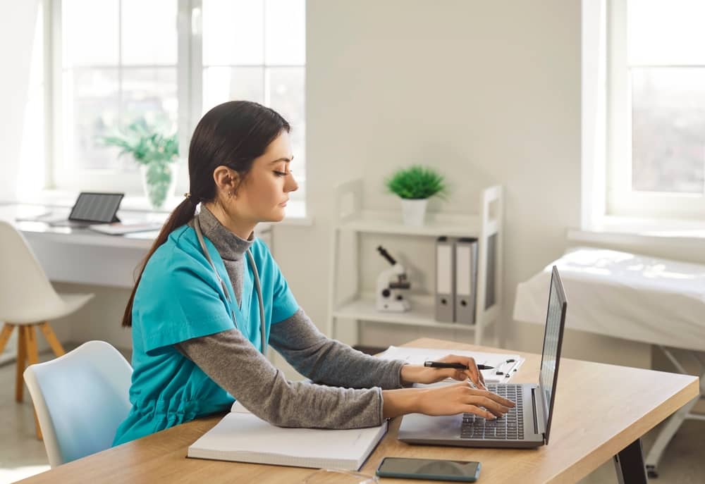 Nurse in teal scrubs working on laptop at desk in bright clinical office