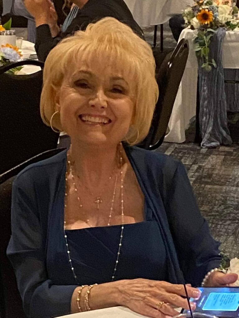 Evelyn, a smiling blonde woman wearing a navy blue dress and delicate layered necklaces, seated at an event