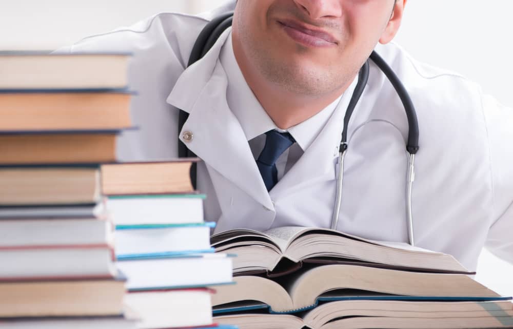 Medical professional in white coat with stethoscope reviewing open medical textbooks and research journals stacked on desk