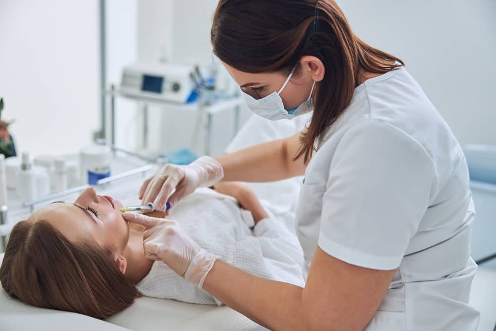 Nurse injector wearing mask and gloves performing facial injectable treatment on female patient in clinical med spa setting