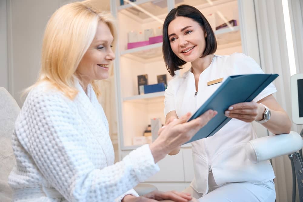 Aesthetic provider in white medical uniform showing digital tablet with treatment information to smiling blonde patient in modern clinic consultation room