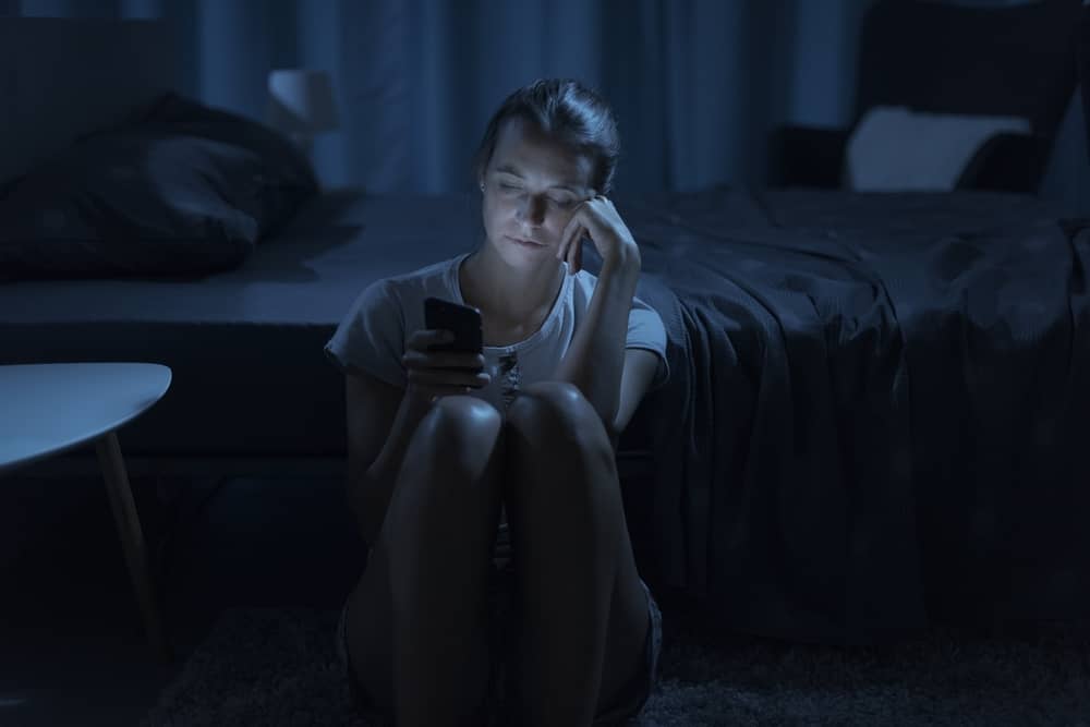 Woman sitting on bedroom floor late at night, face lit by phone screen, searching for answers about facial changes after weight loss
